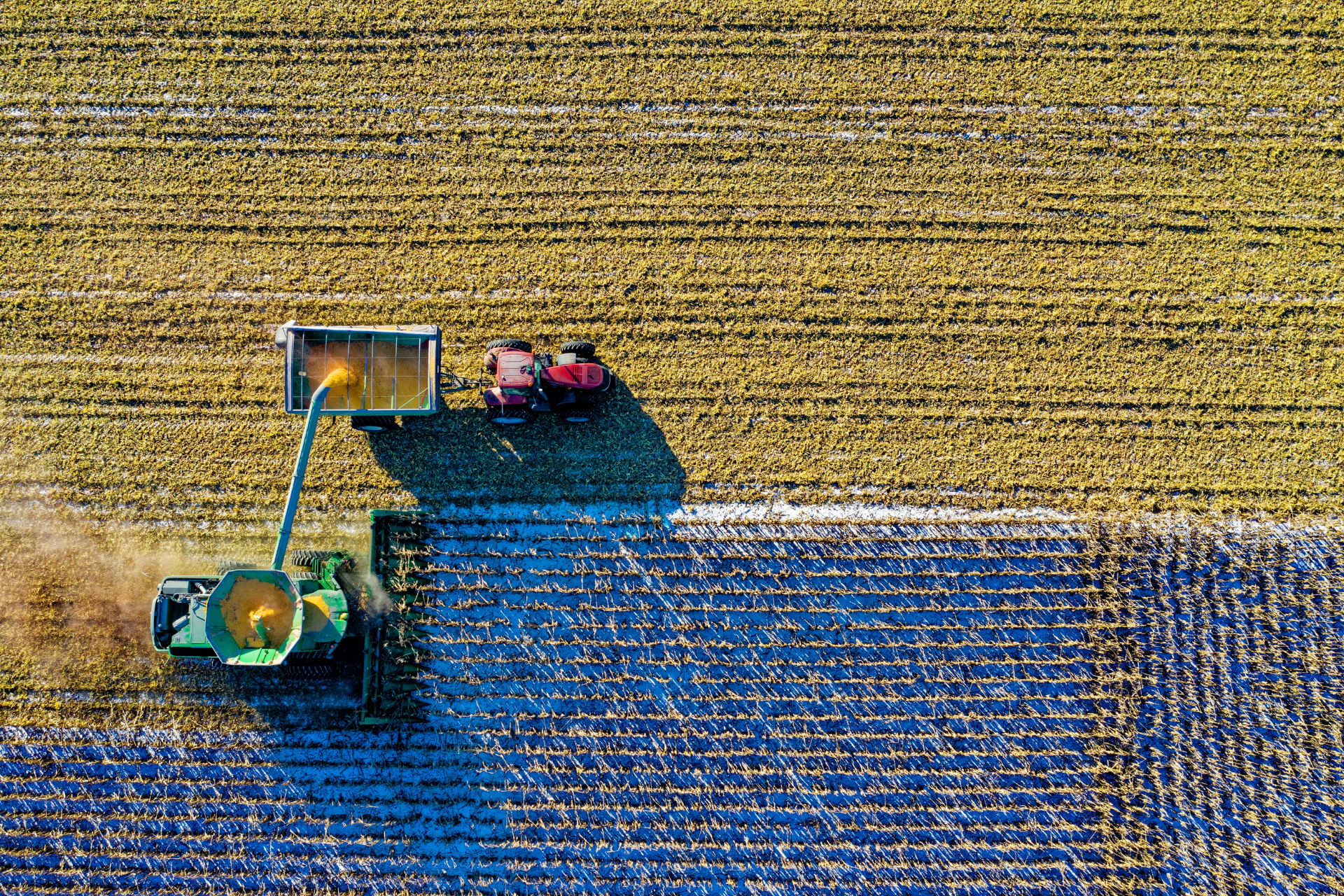 Bird’s eye view of a field in which a harvester is depositing crops into a trailer being pulled by a tractor