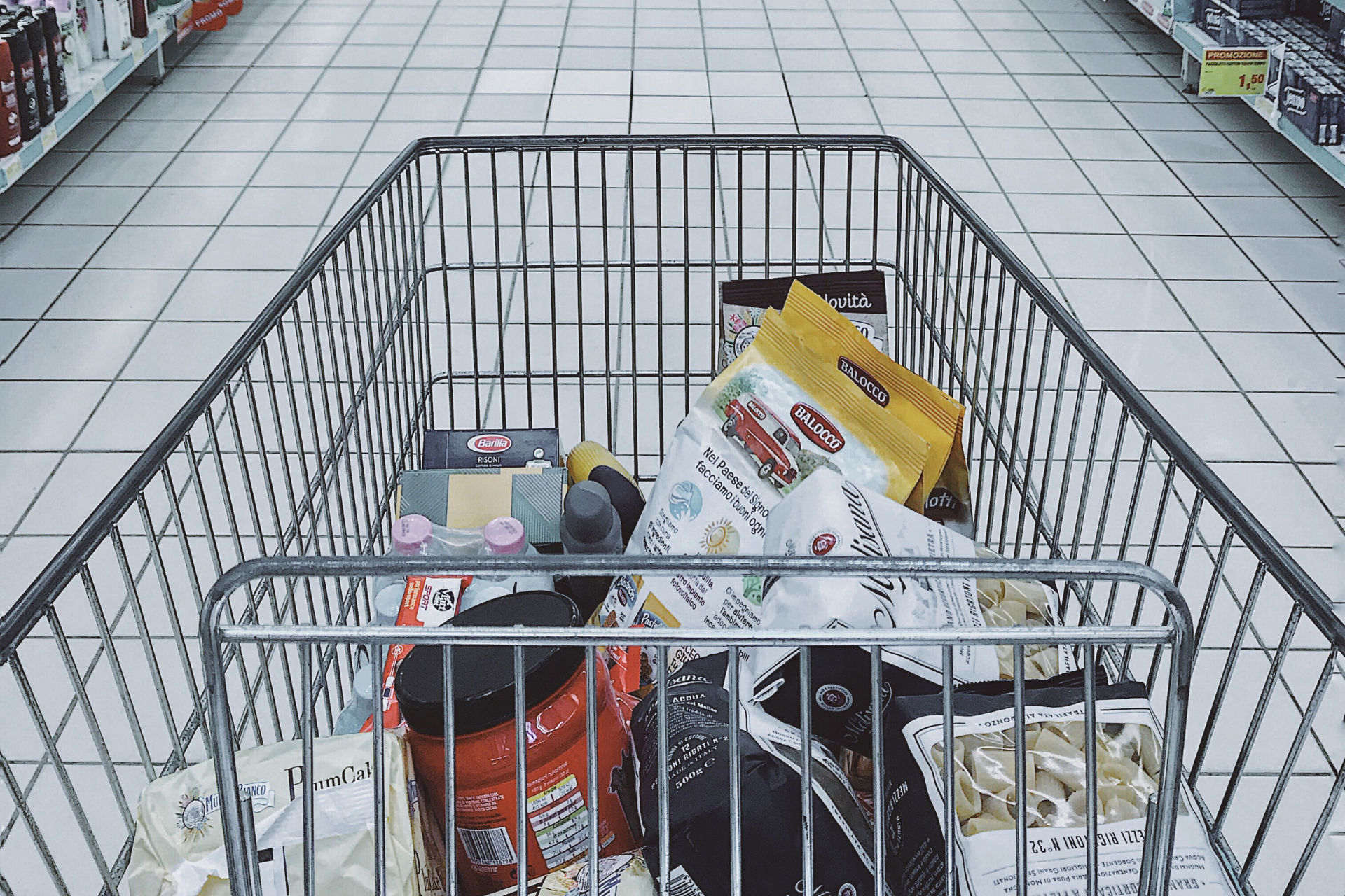  A wire shopping cart filled with assorted groceries, being pushed down a brightly lit grocery store aisle.