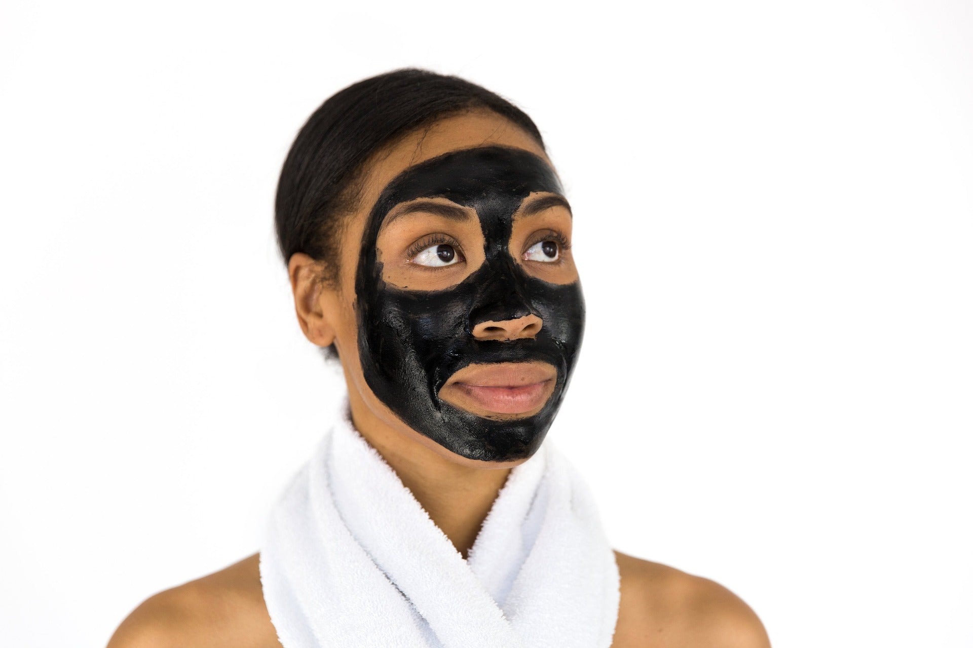 A person with towels around their upper body and neck wears a black cosmetic facemask while holding up the jar of skin treatment cream.