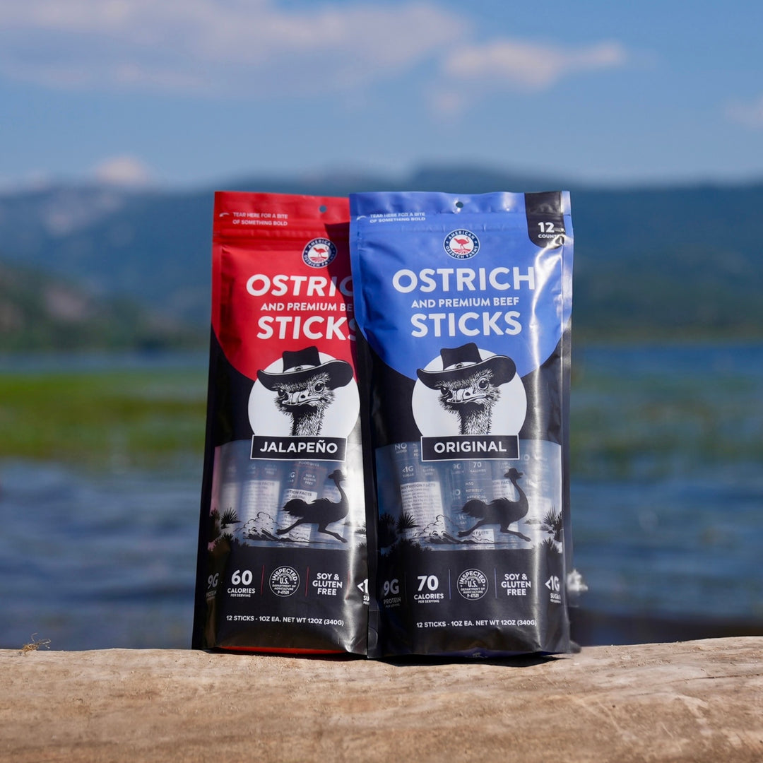 Two packages of Ostrich and Beef Sticks standing upright on a wooden ledge outdoors with a lake and mountains in the background. The left package is red and black labeled Jalapeño, and the right package is blue and black labeled Original.